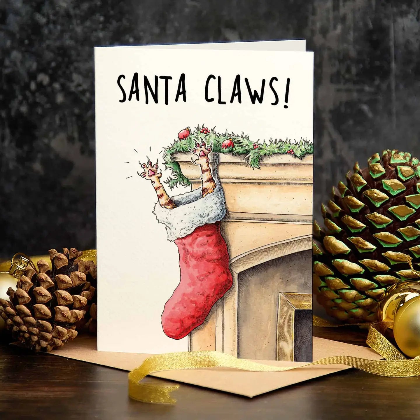 Photograph of a Christmas card with pine cones and other Christmas decorations in the background. The Christmas card shows a decorated mantel with a Christmas sock attached to it from which the front paws with buckled nails of a cat protrude. At the top is the text "Santa Claws!"